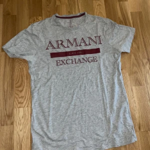 Grå Armani Exchange t-shirt - Snygg grå t-shirt från Armani Exchange med stort vinrött tryck framtill. Klassisk passform med rund hals och korta ärmar. Perfekt för dig som gillar stilrena märkesplagg med en modern touch.