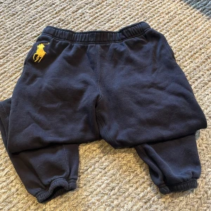 Svarta mjukisbyxor Polo Ralph Lauren - Svarta mjukisbyxor från Polo Ralph Lauren med gul broderad logga på höften. Byxorna har resår i midjan och vid bensluten för skön passform. Tillverkade i mjukt bomullsmaterial, perfekta för chill eller träning.