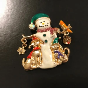 Julbrosch med snögubbe och djur - Superfin brosch med snögubbe, hund och katt i guldton. Snögubben har grön mössa och rosa halsduk, hunden har röd scarf och katten lila scarf samt grön mössa. Dekorerad med små hängen som snöflinga och hjärta. Perfekt accessoar till julens outfits!