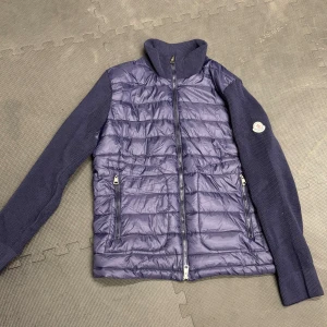 Mörkblå Moncler pufferjacka - Snygg mörkblå pufferjacka från Moncler med stickade ärmar och quiltad front. Jackan har dragkedja framtill, två fickor med dragkedja och klassisk Moncler-logga på ena ärmen. Perfekt för dig som vill ha en stilren och sportig look.