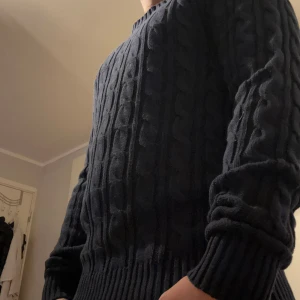 Mörkblå kabelstickad tröja - Säljer en mörkblå kabelstickad tröja med klassiskt mönster och rund hals. Tröjan har långa ärmar och breda ribbade muddar vid ärmslut och nederkant. Perfekt för kyliga dagar och enkel att matcha med jeans eller chinos.