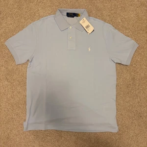 Ljusblå pikétröja från Polo Ralph Lauren - Snygg ljusblå pikétröja från Polo Ralph Lauren i custom slim fit. Tröjan har klassisk krage, korta ärmar och två knappar framtill. Ikoniska broderade loggan i vitt på bröstet. Perfekt för dig som gillar stilrena och fräscha plagg.