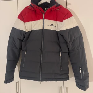 J.lindeberg dunjacka - Snygg pufferjacka från J.Lindeberg med färgblock i rött, vitt och mörkgrått. Jackan har huva, dragkedja framtill och praktiska fickor med dragkedja. Perfekt för kalla dagar och riktigt stilren look.