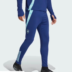 Blå Arsenal träningsbyxor Adidas - Snygga blå träningsbyxor från Adidas med Arsenal-logga på låret och ljusblå detaljer längs sidorna. Byxorna har smal passform, elastisk midja och tre klassiska Adidas-ränder vid vristen. Perfekta för fotboll och sport. Passar även M-L.Pris kan diskuteras 