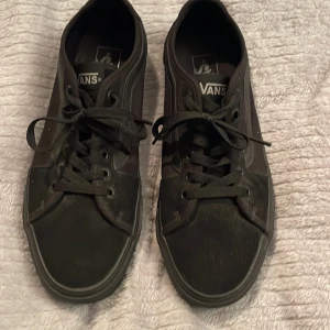 Svarta Vans sneakers storlek 42,5 - Klassiska svarta Vans sneakers med snörning och rund tå. Skorna har en platt sula och är tillverkade i canvas och syntet med Vans logga baktill. Perfekta för dig som gillar streetstyle och enkel design.