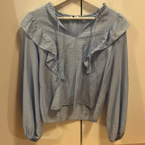 Ljusblå blus med volanger från Kappahl - Supersöt ljusblå blus från Kappahl i storlek XS. Blusen har v-ringning, långa ärmar med resår vid ärmslut och snygga volangdetaljer längs axlarna och framtill. Materialet är lätt och har en skrynklig struktur som ger en cool vibe. Perfekt för dig som gillar romantiska detaljer.💗