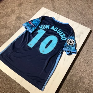 Manchester City Agüero Champions League tröja - Snygg Manchester City bortatröja från Nike med Agüero 10 på ryggen. Mörkblå bas med ljusblå detaljer, tryckta klubb- och sponsorloggor samt Champions League-märke på ärmen. Tillverkad i lätt och ventilerande polyester.
