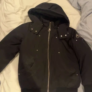 Svart pufferjacka Moose Knuckles - Svart pufferjacka från Moose Knuckles med huva, silvriga nitar och dragkedja framtill. Jackan har logga på ärmen och broderad hockeymotiv på insidan. Snygg och varm modell med coola detaljer och ribbade muddar.