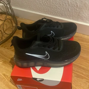 Svarta Nike sneakers med luftig sula - Säljer ett par svarta Nike sneakers med vit swoosh på sidan och transparent, luftig sula. Skorna har snörning och är tillverkade i mesh och syntetmaterial för extra komfort. Perfekta för dig som gillar sportig stil och vill ha något som sticker ut.