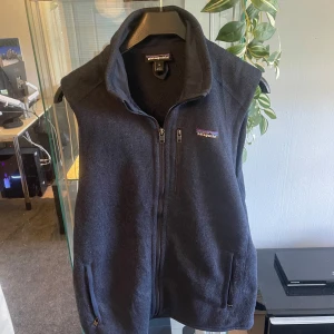 Patagonia väst - Säljer en mörkblå fleeceväst från Patagonia med hög krage och dragkedja framtill. Västen har två fickor med dragkedja och en liten broderad Patagonia-logga på bröstet. Perfekt att slänga över en hoodie eller långärmad tröja.