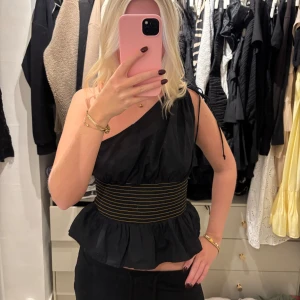 Svart oneshoulder topp med volang - Säljer en svart oneshoulder topp med bred, elastisk midjedetalj i svart och gul/guld. Toppen har volang nedtill. Bra skick!