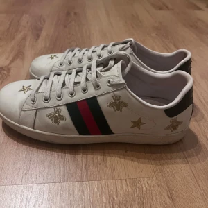 Gucci Ace sneakers 41 - Gucci sneakers i vitt skinn med klassiska gröna och röda ränder på sidorna. Skorna har broderade guldiga bin och stjärnor samt svart detalj med Gucci-logga på hälen. Platt sula och snörning framtill. Perfekta för dig som vill sticka ut med lyxig streetstyle.
