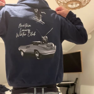 Montévas Winter Luxury Club hoodie - Snygg mörkblå hoodie från Monteira med tryck på ryggen som visar en skidåkare och en retro bil samt texten 'Montéva Luxury Winter Club'. Tröjan har huva och långa ärmar, perfekt för dig som gillar coola vintermotiv och streetwear.