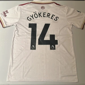 Arsenal Gyökeres 14 bortatröja Adidas - Snygg Arsenal bortatröja med Gyökeres 14-tryck. Vit bas med vinröda detaljer, broderat klubbmärke och Adidas-logga. Klassisk krage med ränder på axlarna och subtilt geometriskt mönster i tyget. Tillverkad i lätt och ventilerande polyester (AEROREADY). Replika, endast handtvätt gäller!