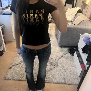 Svart croppad topp med guldtext - Säljer en svart croppad topp med korta ärmar och glittrig guldtext 'PARIS' framtill. T-shirten har en slim passform.