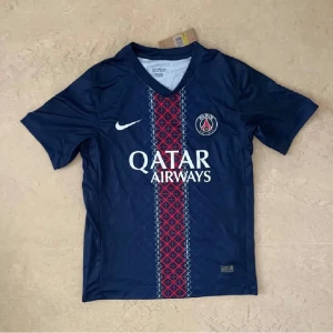 PSG blå fotbollströja Nike - Snygg Paris Saint-Germain fotbollströja från Nike i mörkblått med rött och vitt geometriskt mönster längs mitten. Tröjan har korta ärmar, V-ringad hals och Qatar Airways tryck på bröstet. Perfekt för dig som älskar fotboll och PSG.