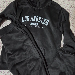 Svart Träningsset med Los Angeles  - Svart hoodie med vit Los Angeles-tryck på bröstet och snörning vid huvan. Tröjan har en stor magficka och långa ärmar. Materialet känns mjukt och är troligen bomull eller polyester, perfekt för chill dagar. Snygg och enkel streetstil.