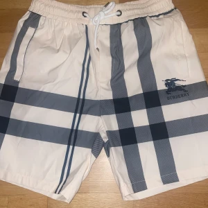 Burberry rutig badshorts med snörning - Snygga badshorts från Burberry i klassiskt rutigt mönster i vitt och mörkblått. Shortsen har elastisk midja med vit snörning, sidofickor och Burberry-logga på ena benet. Perfekta för sommaren och strandhäng.  (STRL M)