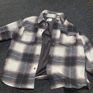Rutig overshirt från H&M i grått - Säljer en snygg rutig jacka från H&M i grå, vit och svart ton. Jackan har två stora bröstfickor med lock, knappar framtill och klassisk krage. Materialet är mjukt och känns lite som ull, perfekt för lager på lager. Passformen är loose och den har långa ärmar. Passar även för lite kalla blåsiga dagar. Använd fåtal gånger köpte för 599 säljer för 450, pris går att diskuteras