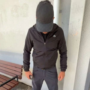 Lacoste vindjacka  - ”JACKAN ÄR ÄKTA” köpte den för 2000,, Svart vindjacka från Lacoste Sport med huva och dragkedja framtill. Jackan har det klassiska gröna krokodilmärket på bröstet och meshfoder på insidan för extra komfort. Regular fit och stilren design som funkar till streetwear. Perfekt för dig som gillar sportiga vibes.
