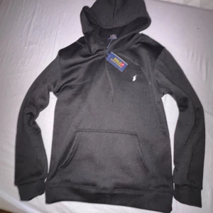 Svart Ralph lauren hoodie - Svart hoodie från Polo Ralph Lauren med klassisk huva och känguruficka. Tröjan har en vit broderad logga på bröstet och snörning vid halsen. Mjuk bomullskvalitet och ribbade muddar vid ärmslut och nederkant. Perfekt för chill dagar.