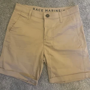 Beige shorts från Race Marine XS - Snygga beiga shorts från Race Marine i klassisk chino-stil. De har normal passform, bälteshällor, framfickor och två bakfickor. Perfekta för varma dagar och lätta att matcha med det mesta. Tillverkade i mjuk bomull för en skön känsla.