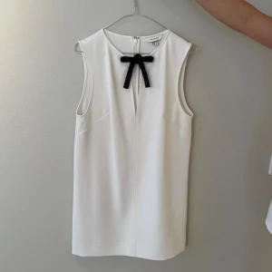 Vit ärmlös kortklänning med rosett - Snygg vit kortklänning från H&M med rak passform och ärmlös design. Klänningen har en svart rosett vid halsen som ger en chic touch. Tillverkad i mjuk polyester och har en diskret dragkedja bak. Perfekt för dig som gillar stilrena och moderna plagg.