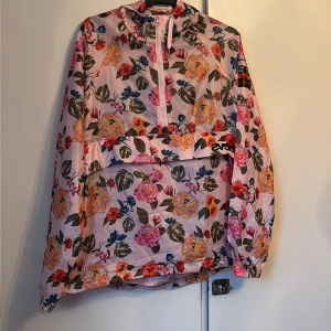 Blommig vindjacka från Svea - Säljer en riktigt snygg vindjacka från Svea i medium. Jackan har ett blommigt mönster i rosa, orange, blå och gröna toner, med halv dragkedja framtill och stor ficka. Perfekt för dig som vill sticka ut med färg och mönster.