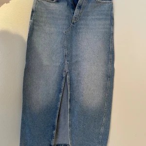 Lång jeanskjol med slits från MNG - Snygg lång jeanskjol från MNG i klassisk blå denim. Kjolen har en rak passform, hög midja, slits framtill och två bakfickor. Perfekt för dig som gillar en stilren och tidlös look. Materialet är bomull och kjolen har dragkedja och knapp framtill.