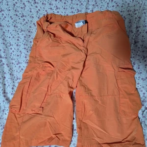 Orange cargo shorts Knot So Bad M - Säljer ett par orangea cargo shorts från Knot So Bad i storlek M. De har flera praktiska fickor på sidorna och bak, samt knapp och dragkedja framtill. Materialet är lätt och skönt, perfekt för varma dagar. Midjan är elastisk för extra komfort.