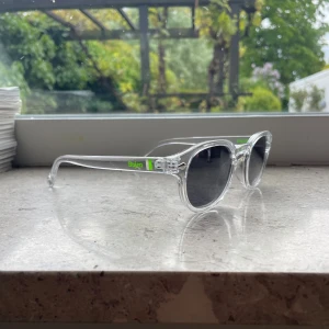Bajen transparenta solglasögon (Limited edition)  - Snygga solglasögon från Moritz med transparent båge och svarta runda linser. På skalmen finns en grön Bajen-logga som ger extra attityd. Glasögonen har en modern och stilren look som passar perfekt för soliga dagar. Sprillans nya!! 