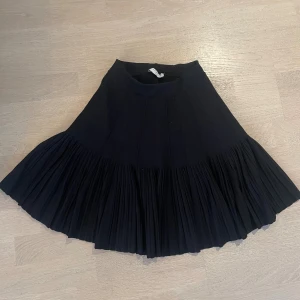 Plisserad svart kjol från Sandro - Supersnygg svart kjol från Sandro med plisserad nederdel och bred midja. Kjolen har en kort modell och är tillverkad i ett stickat material som ger en lyxig känsla. Perfekt för dig som vill ha en klassisk men trendig look.