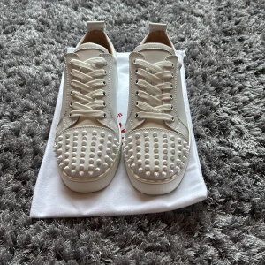 Christian Louboutin vita sneakers med nitar - Säljer ett par sjukt snygga vita sneakers från Christian Louboutin med coola vita nitar på tån och klassisk röd sula. Skorna har snörning och är gjorda i skinn med rund tå och platt sula. Perfekt för dig som vill sticka ut med exklusiv stil.