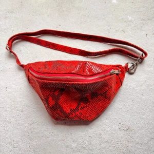 Röd magväska i skinn från Vimoda - Snygg röd magväska från Vimoda med coolt ormmönster och justerbar rem. Väskan är gjord i skinn och har en dragkedja upptill samt ett ljust innerfoder med extra fack. Perfekt för att bära det viktigaste och ge din outfit en edgy vibe.