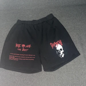 Svarta shorts med dödskalle från Romwe - Svarta shorts från Romwe i storlek S med elastisk midja. Framsidan har tryck med texten 'WE ARE THE BEST' och 'SHADOW' samt en stor vit dödskalle. Röd och vit text ger en edgy vibe. Materialet är mjuk bomull, perfekt för chill dagar.