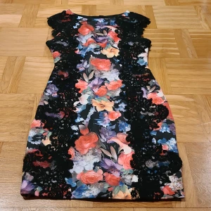 Blommig kortklänning från Joseph Ribkoff - Supersnygg kortklänning från Joseph Ribkoff med färgstarkt blommönster i rött, blått, lila och vitt. Klänningen har svarta spetsdetaljer längs sidorna och axlarna, figurnära passform och är ärmlös. Perfekt för dig som vill sticka ut med en unik och färgglad look.