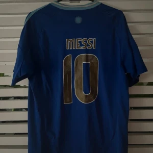 Blå Argentina Messi 10 Adidas tröja - Säljer en blå Argentina fotbollströja från Adidas med Messi och nummer 10 på ryggen. Tröjan har ljusblå detaljer, AFA-emblem på bröstet och klassiska Adidas-ränder på axlarna. Perfekt för dig som älskar fotboll och Messi.