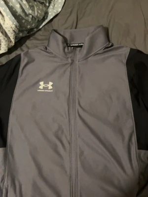 Grå träningströja  från Under Armour - Säljer en grå träningströja  från Under Armour med svarta ärmar och hel dragkedja framtill. Jackan har hög krage och logga på bröstet. Perfekt för träning eller uppvärmning. Materialet är troligtvis polyester för en sportig känsla.