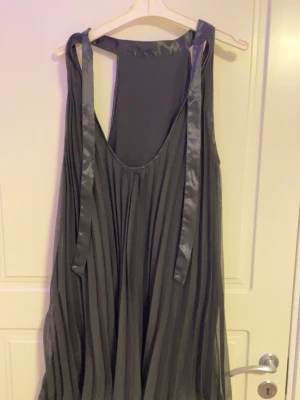 Mörkgrå omlottklänning med band - Säljer en mörkgrå omlottklänning med breda axelband i satin och plisserad kjol. Klänningen har ett flowy fall och knytband vid axlarna för en cool touch. Perfekt för dig som gillar en avslappnad men ändå chic stil.