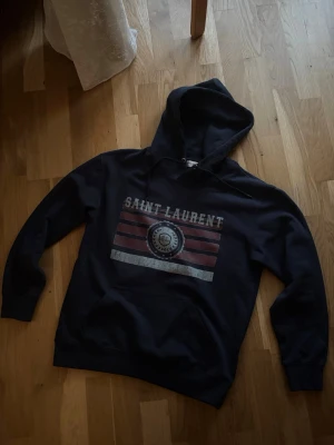 Mörkblå Saint Laurent hoodie  - Säljer min mörkblåa Saint Laurent hoodie. Hoodien är i mycket bra skick och är knappast använd. Den är väldigt stilig samt bekväm. Ställ gärna frågor och ge prisförslag!