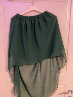 Grön asymmetrisk långkjol - Säljer en flowy grön långkjol med asymmetrisk fåll och resår i midjan. Kjolen har ett tunt, skirt material som ger ett snyggt fall och passar perfekt till en trendig och avslappnad stil.