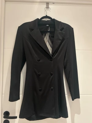 Svart kavaj med glittriga detaljer - Säljer en svart kavaj med dubbelknäppning och långa ärmar. Baksidan har snygga glittriga fransdetaljer i silver som ger en unik look. Perfekt för dig som vill sticka ut! Kavajen har en figurnära passform och klassisk krage.