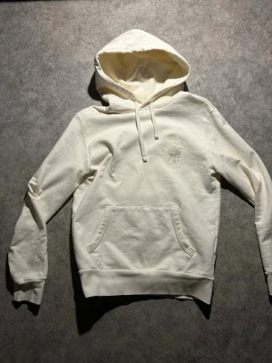 Vit/kräm hoodie från Polo Ralph Lauren - Snygg vit hoodie från Polo Ralph Lauren med klassisk broderad logga på bröstet i samma färg som tyget. Hoodien har huva med dragsko, magficka och ribbade muddar vid ärmar och nederkant. Tillverkad i mjuk bomull för maximal komfort. Köpt från Ralph laurens hemsida självklart äkta.