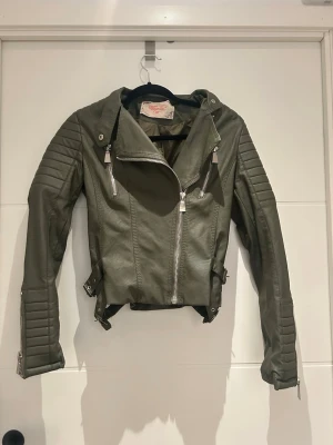 Olivgrön biker skinnjacka med dragkedjor - Snygg olivgrön biker skinnjacka från voyelles med coola detaljer som dragkedjor, quiltade partier på ärmarna och justerbara spännen i midjan. Jackan har en croppad passform och långa ärmar med dragkedjor vid ärmsluten. Perfekt för dig som vill ha en edgy look.