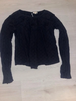 Mörkblå blus med prickar från Gina Tricot - Snygg mörkblå blus från Gina Tricot med diskreta prickar i tyget och volangdetaljer vid axlarna. Blusen har rund halsringning och långa ärmar med lätt rynkade muddar. Perfekt för en trendig och avslappnad look.