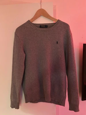 Grå stickad tröja Polo Ralph Lauren - Klassisk grå stickad tröja från Polo Ralph Lauren med rund halsringning och diskret broderad logga på bröstet. Tröjan har ribbade muddar vid ärmslut och nederkant, perfekt för en stilren och enkel look.