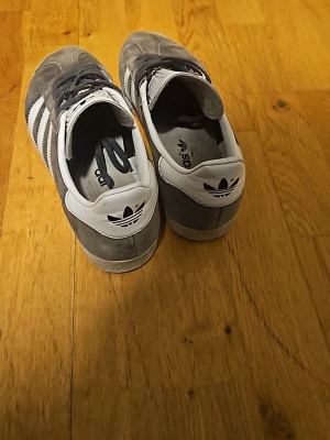 Grå Adidas Gazelle sneakers mocka - Säljer ett par grå Adidas Gazelle sneakers i mocka med vita detaljer och klassiska tre ränder på sidan. Skorna har snörning, vit plös och häl med Adidas-logga. Perfekta för dig som gillar stilrena och tidlösa sneakers. 8/10 skick