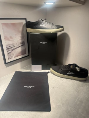 Svarta Saint Laurent sneakers - Säljer ett par svarta Saint Laurent sneakers med vit sula och handskriven logga på sidan. Skorna har snörning och är tillverkade i canvas med klassisk låg profil. Perfekta för dig som gillar stilrena och exklusiva sneakers. Pris kan diskuteras!