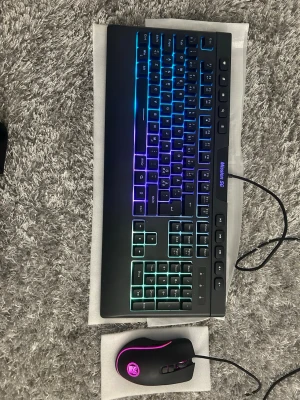 Gamingtangentbord och mus med RGB-belysning - Säljer ett svart gamingtangentbord med färgglad RGB-belysning och tillhörande svart gamingmus med rosa LED-ljus. Båda är kabelanslutna och har en modern design, perfekt för gaming eller datoranvändning. Tangentbordet har numerisk del och mediaknappar.