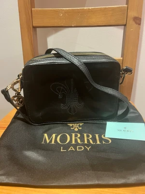 Svart axelväska från Morris Lady - Snygg svart axelväska från Morris Lady i skinn med guldfärgade metalldetaljer och justerbar axelrem. Väskan har ett elegant mönster framtill och dragkedja upptill. Perfekt för dig som vill ha en stilren och trendig accessoar.